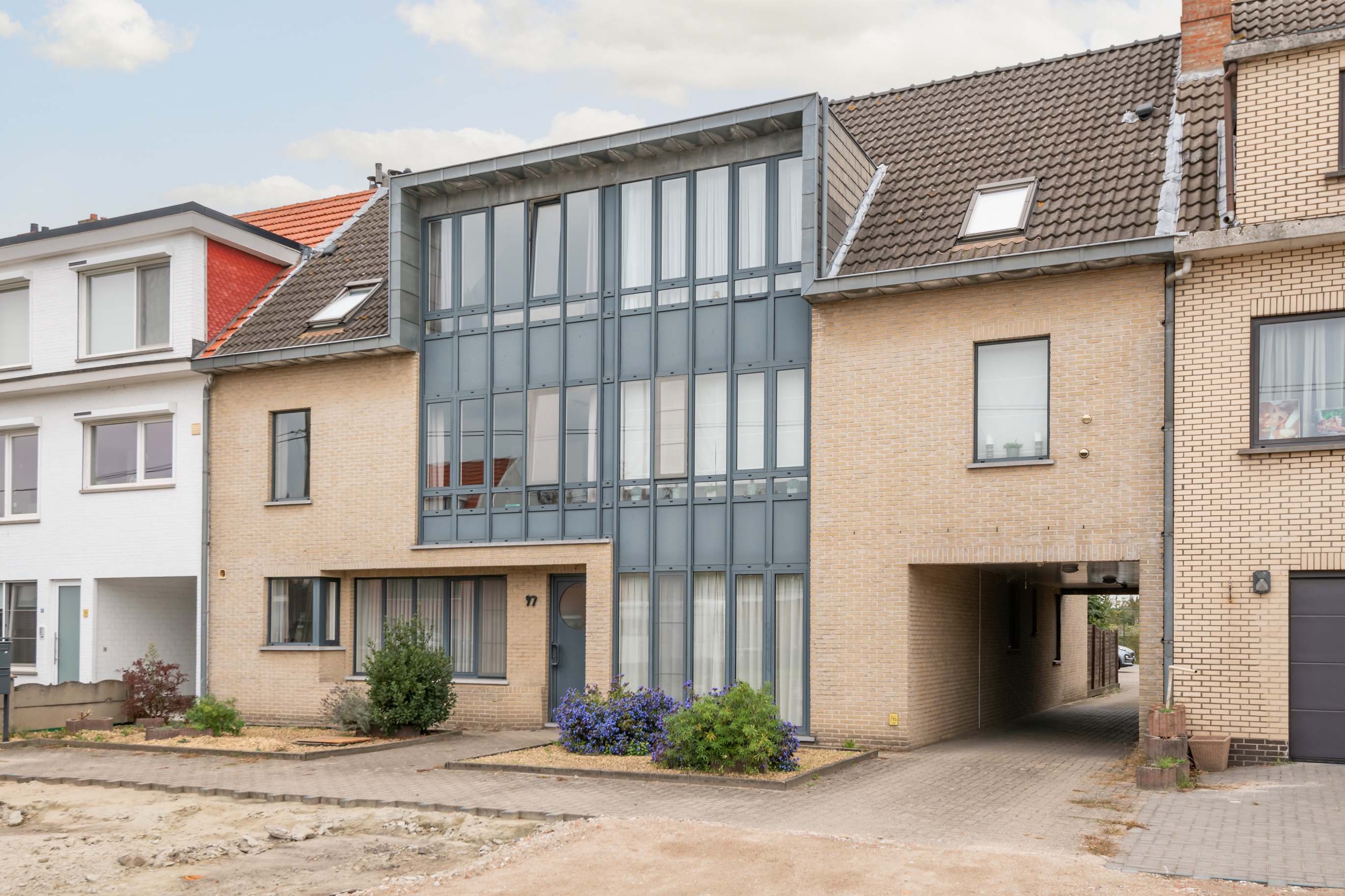 Modern beige brick house with a large grey glass facade, a covered driveway, and small front garden with purple flowers. [AI-generated text]