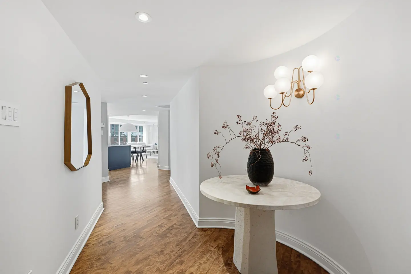Bright hallway with wood floors, white walls, and recessed lighting. A table with a vase and a gold-framed mirror add decor.