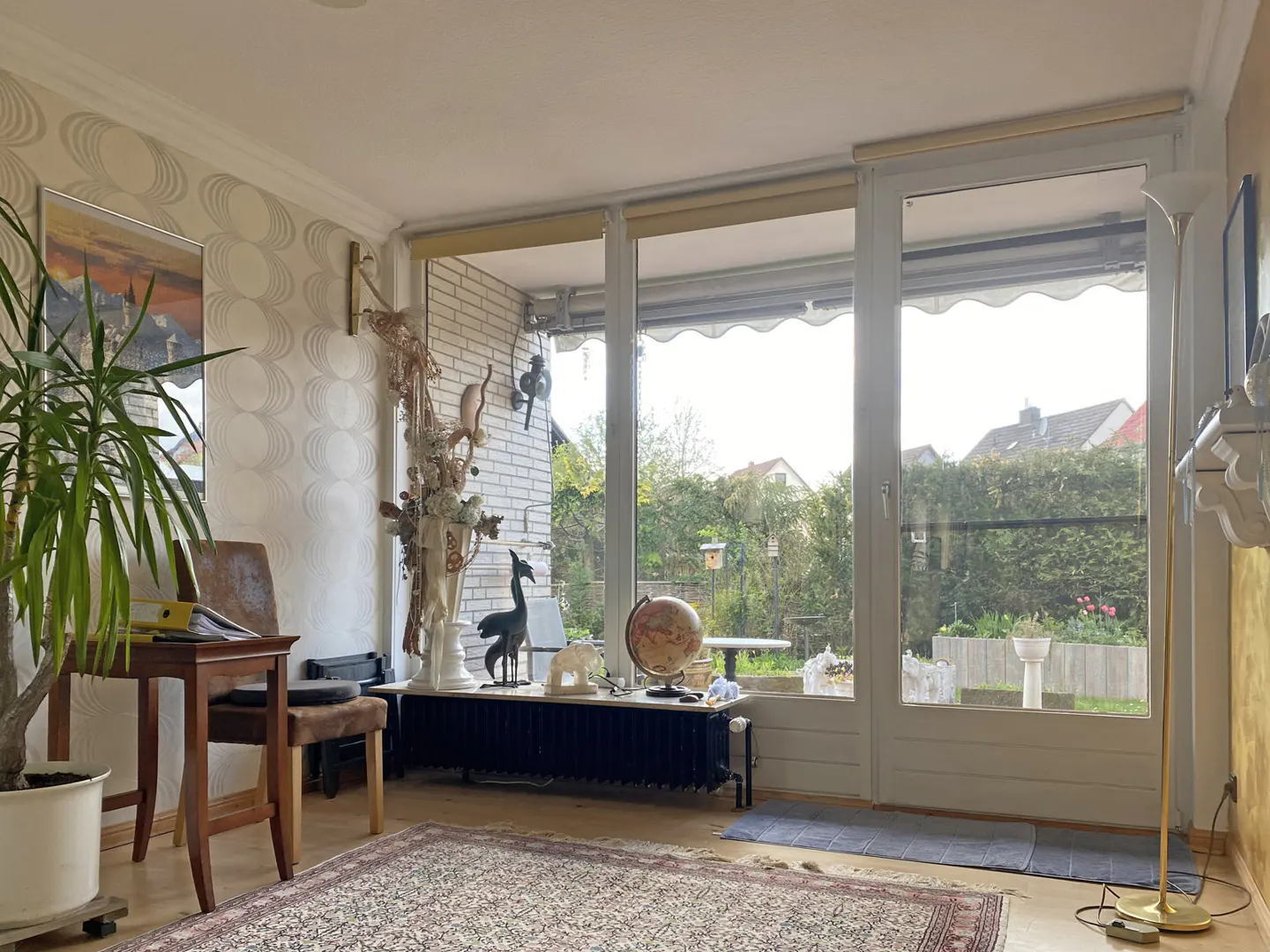 Living room with patterned wallpaper, a wooden desk, and a large window overlooking a green garden. A globe and decorative items sit on a radiator.