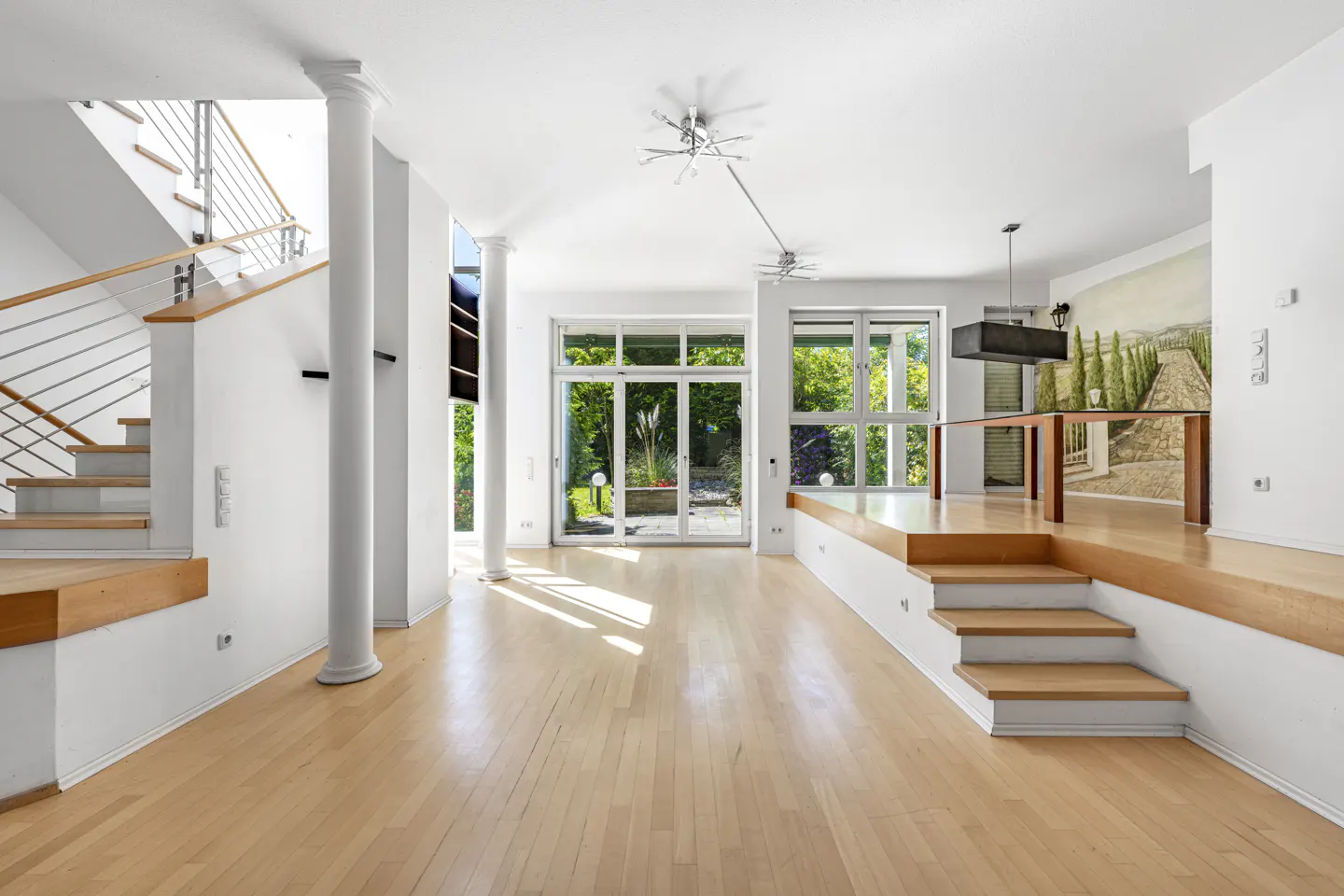 Bright, open-plan living area with light wood floors, a staircase, a raised dining platform, and glass doors to a garden.