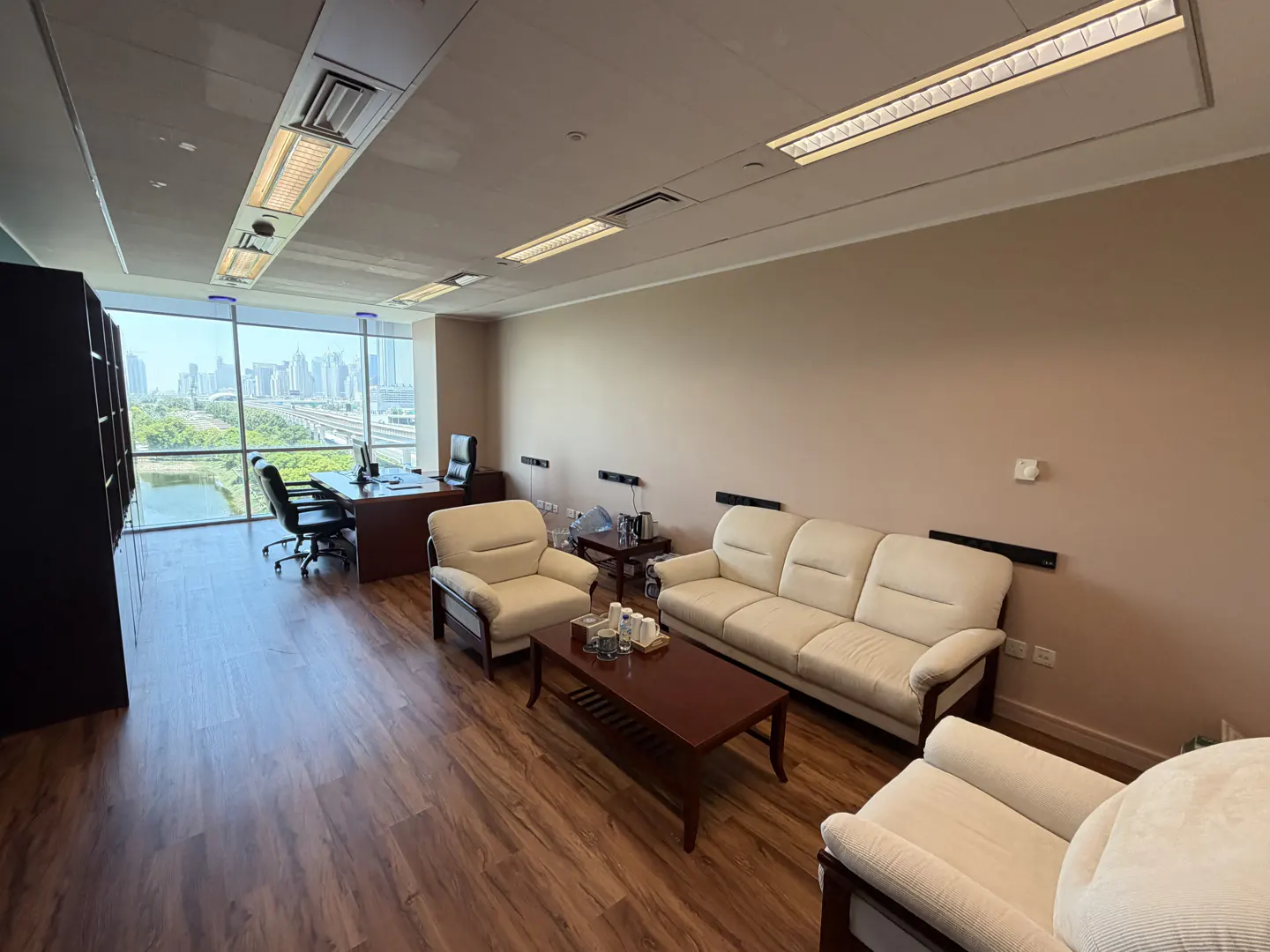 Office interior with wood floors, cream sofa and chairs, and a desk with a city view through a large window.