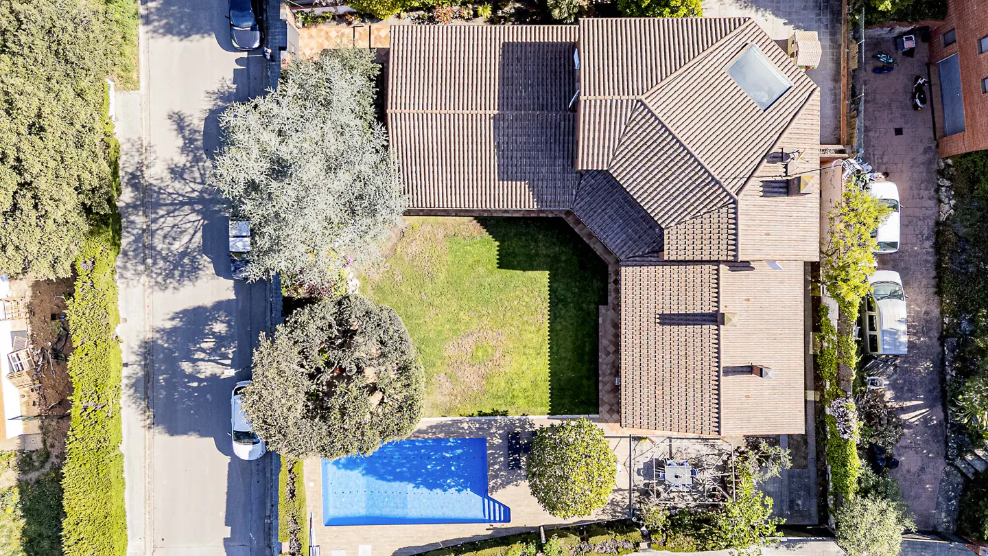 Aerial view of a house with a brown tile roof, green lawn, and blue pool. Trees and cars surround the property.
