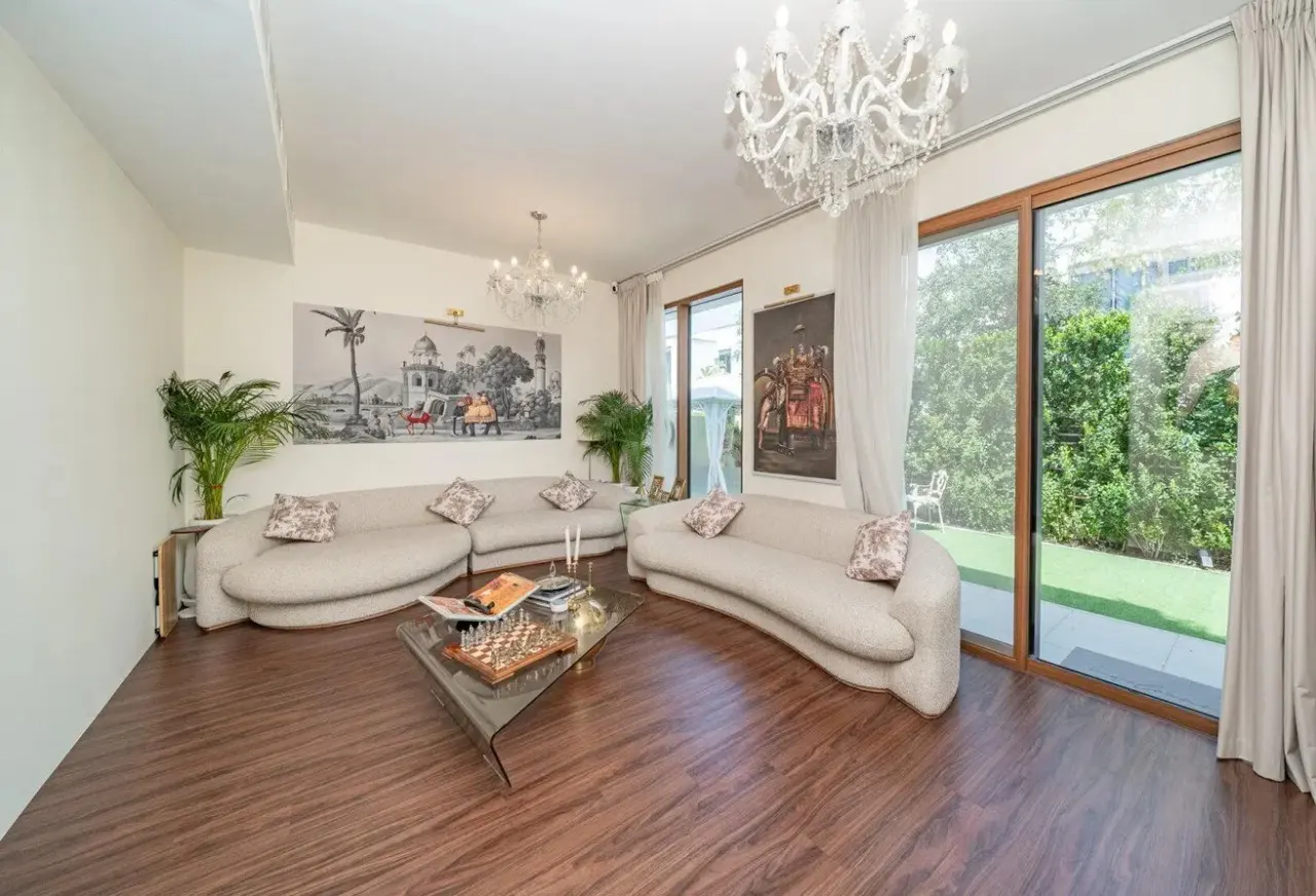 Living room with curved sofas, glass table with chess set, wood floors, and large windows overlooking a green yard. Chandeliers hang from the ceiling.