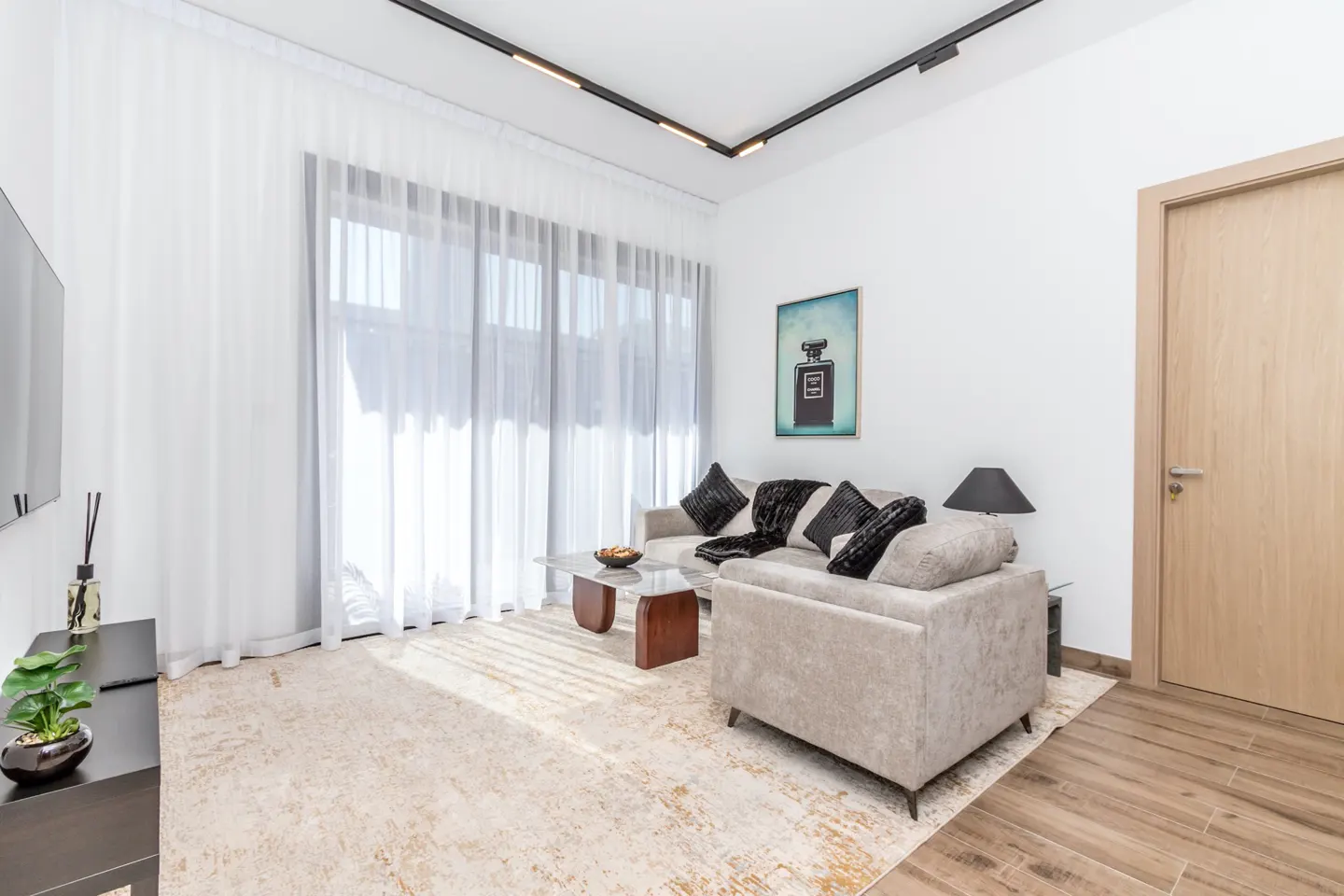 Bright living room with beige sofa, black pillows, and a glass coffee table on a patterned rug. White walls, sheer curtains, and a perfume bottle art piece.
