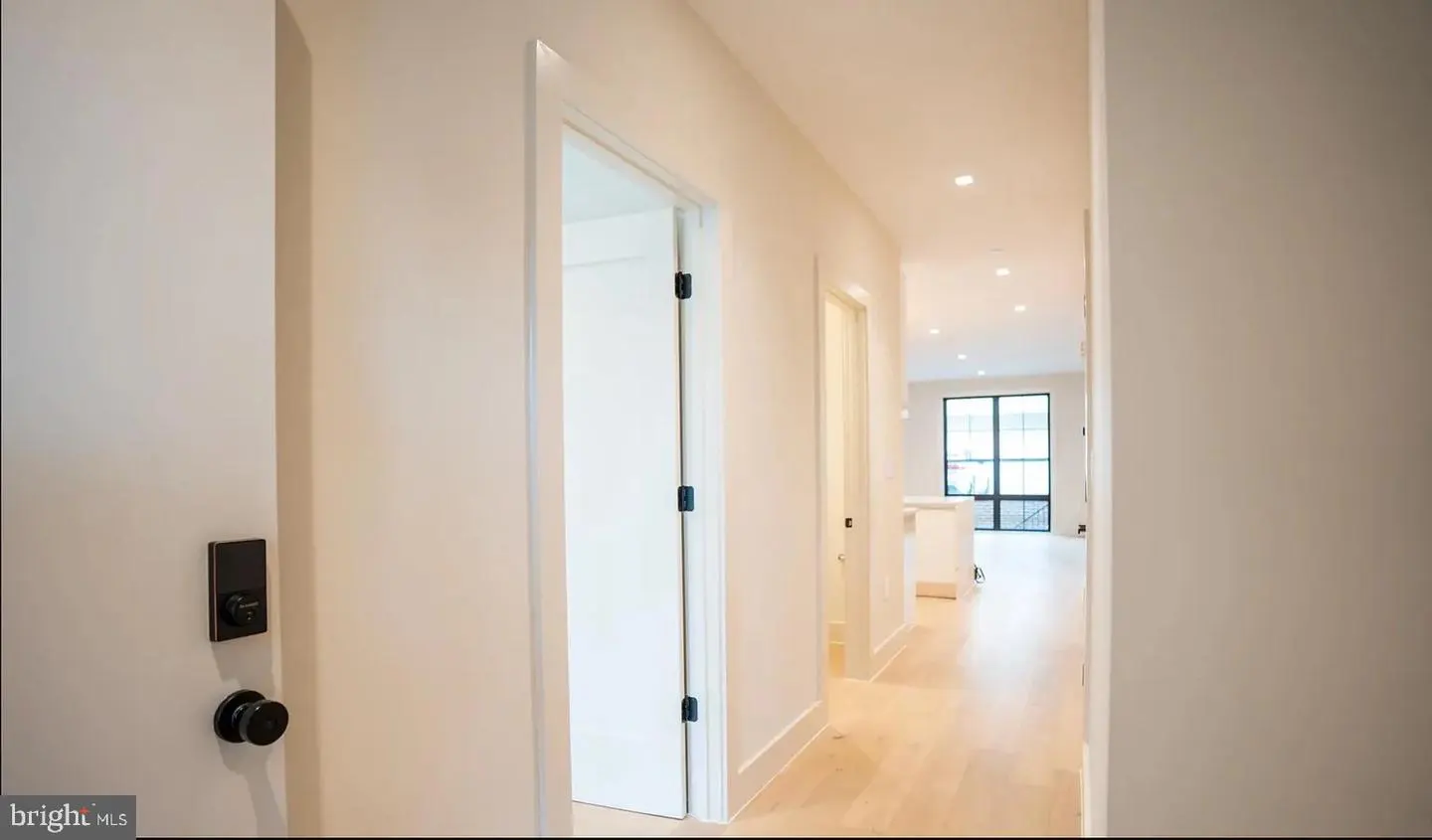 Hallway view with light wood floors, white walls, and recessed lighting. Open doorways lead to a bright living space with a large window.