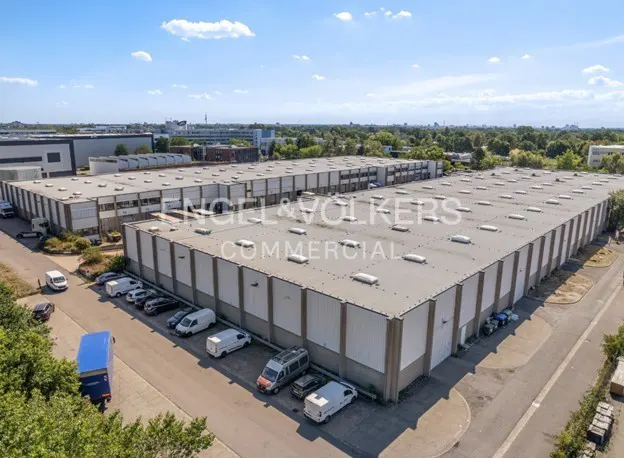 Aerial view of a large, gray commercial building with white accents, surrounded by parked cars and greenery. "Engel & Volkers Commercial" is overlaid.
