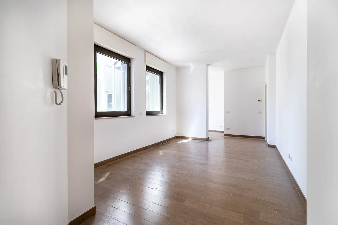 Bright, empty room with white walls, wood floors, and two windows with dark frames. An intercom is mounted on the wall.
