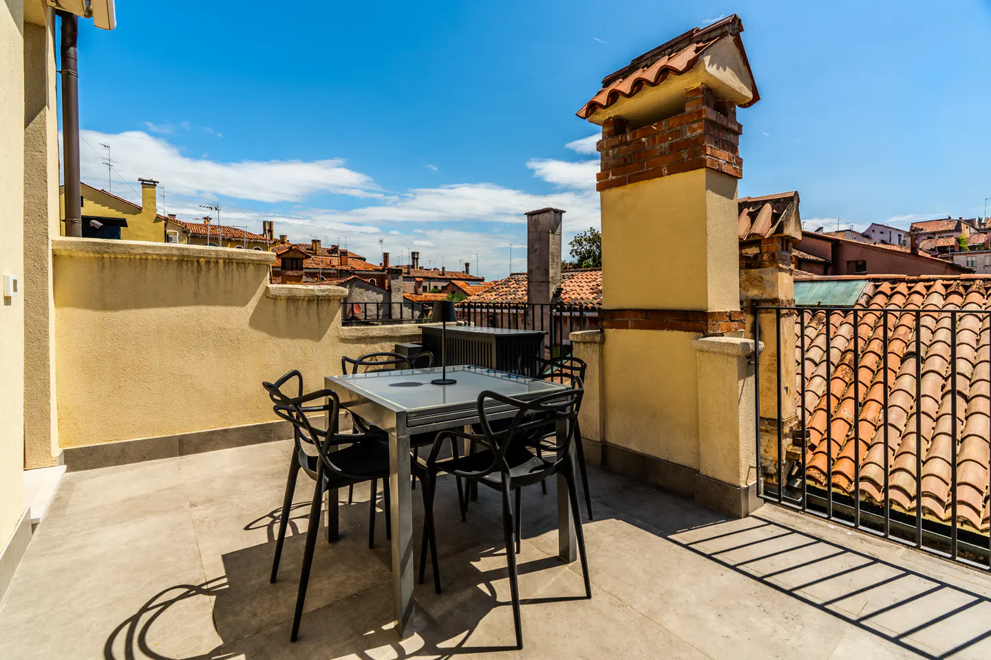 Rooftop terrace with a glass table and black chairs, overlooking a scenic cityscape with terracotta tiled roofs.