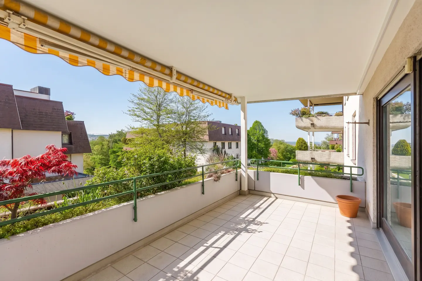Spacious balcony with white tiled floor, yellow striped awning, green railing, and a view of trees and neighboring houses.