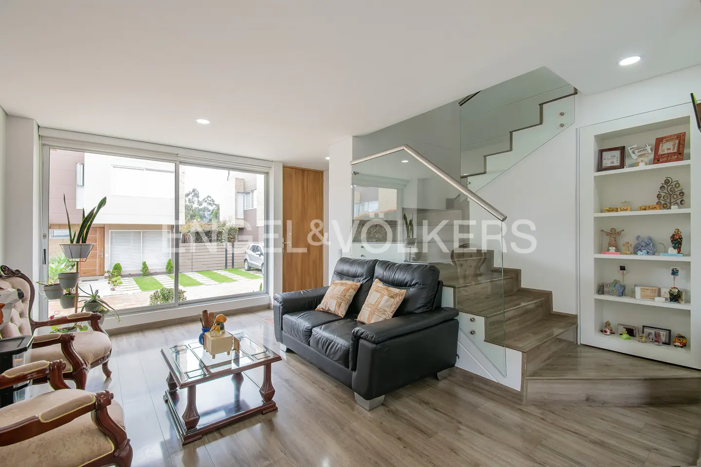 Modern living room with a black leather sofa, glass coffee table, wood floors, and a staircase with a glass railing.