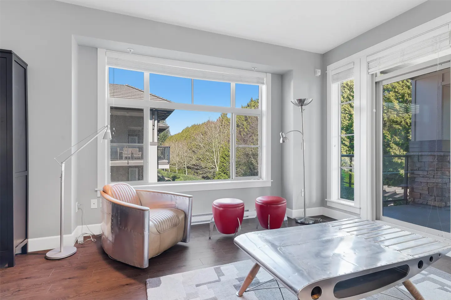 Living room with a metal chair, red stools, and a metal coffee table. A large window overlooks trees and a building.