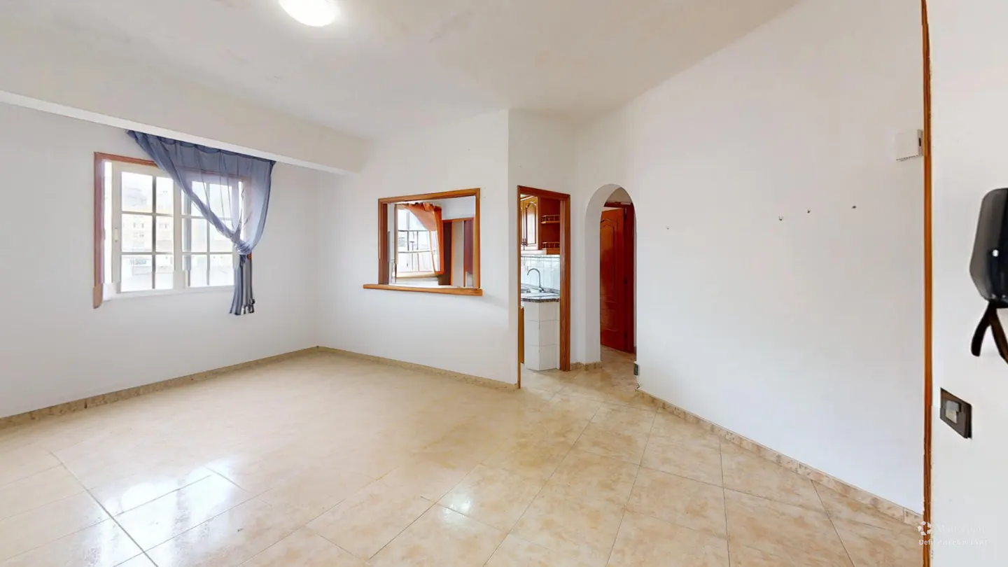 Bright, empty room with white walls, tan tile floor, and a window with blue curtains. A kitchen is visible through a doorway.