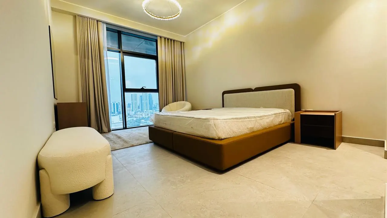 Bedroom with a brown bed, white mattress, nightstand, and a large window with city views. Beige walls and curtains. Modern chandelier.