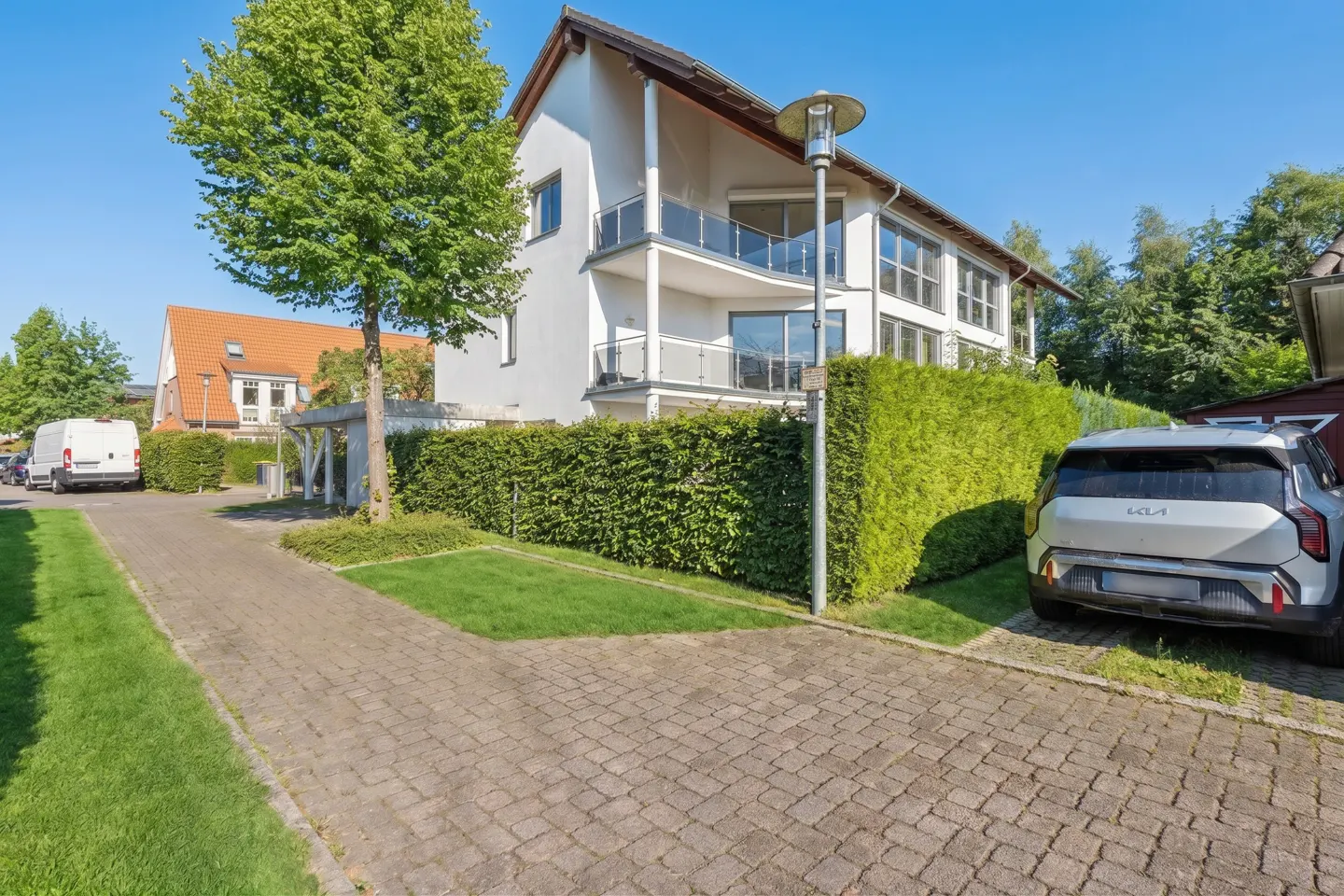 Exterior view of a modern white house with balconies, green hedges, and a silver car parked in the driveway.