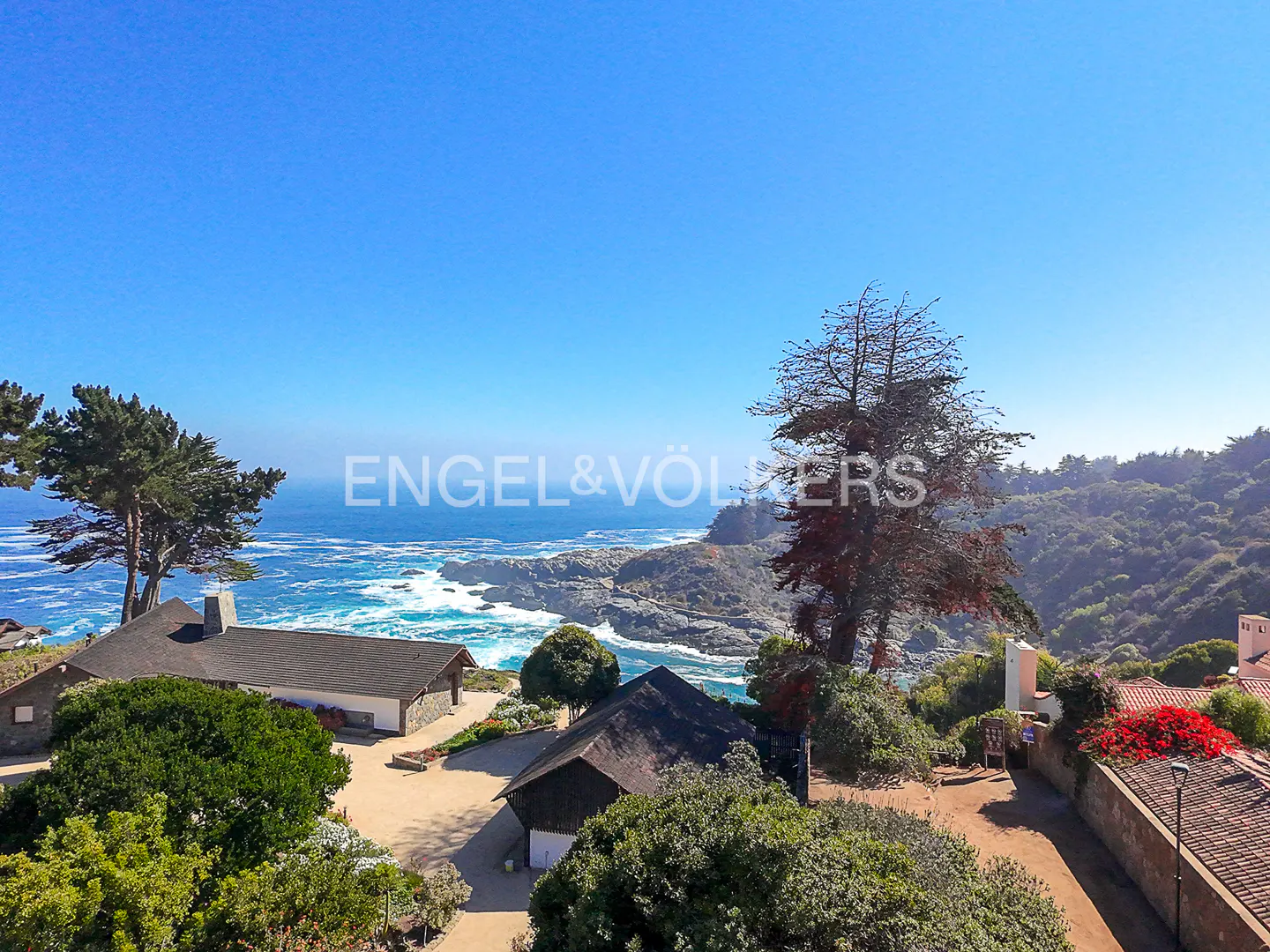 Coastal property with ocean view. Houses with dark roofs, lush greenery, and rocky shoreline under a clear blue sky.