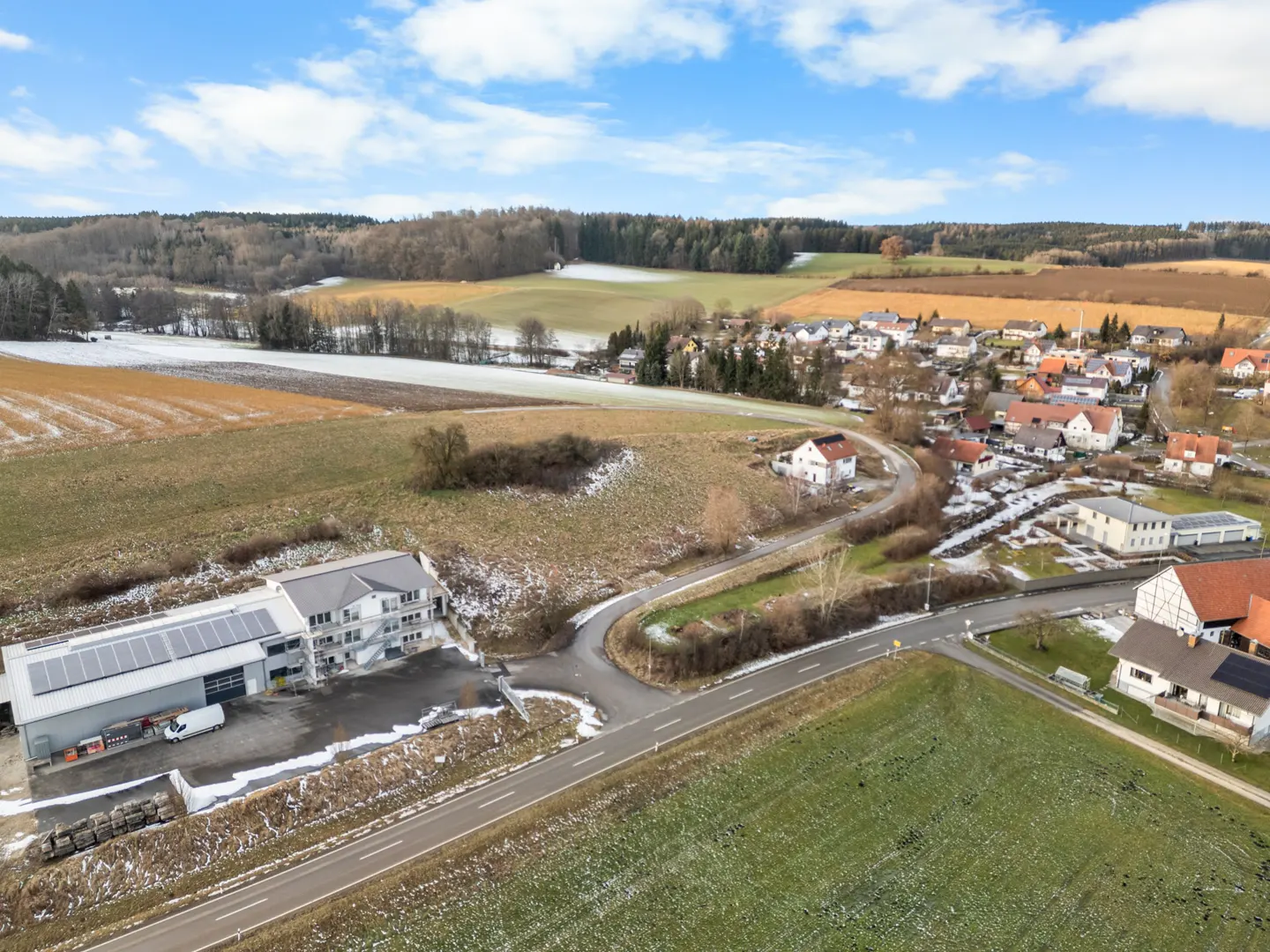 Aerial view of a rural landscape with fields, a village, and a modern building with solar panels on its roof. Patches of snow are visible.