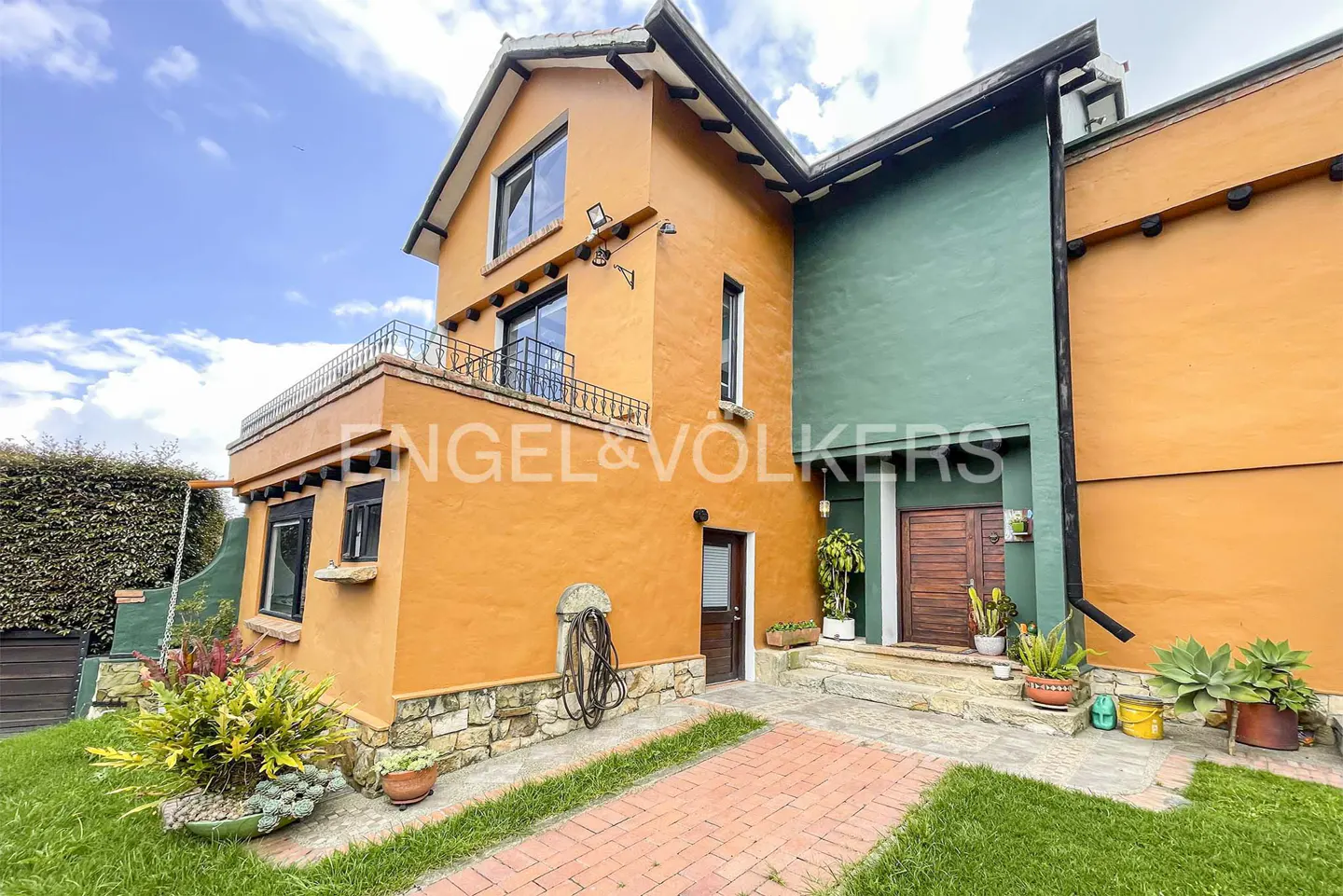 Two-story house with orange and green walls, a brick path, and a lawn. The Engel & Volkers logo is visible on the orange wall.