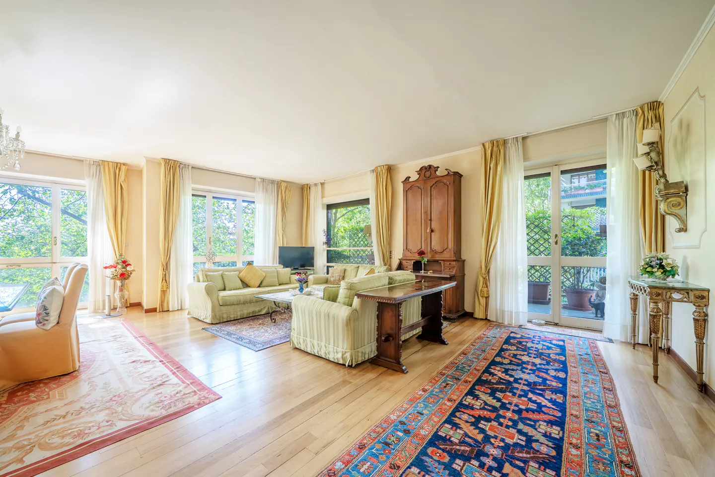 Bright living room with hardwood floors, sofas, and large windows with yellow curtains. A blue patterned rug adds color to the space.