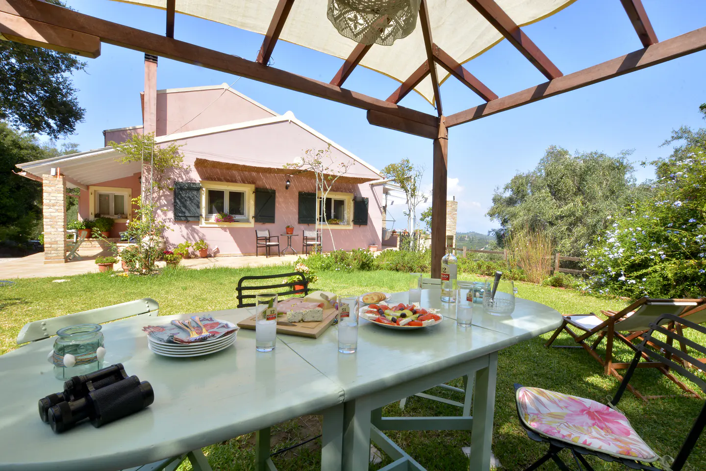 Outdoor dining area with a light green table set for a meal, featuring plates, glasses, and food. A pink house with green shutters is in the background, under a blue sky.