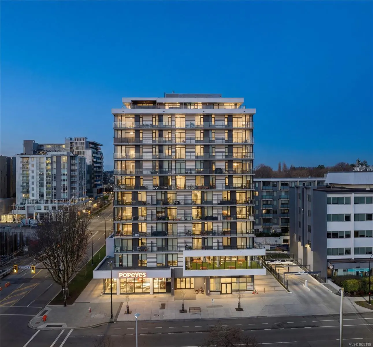 Exterior shot of a modern, multi-story apartment building with a Popeyes restaurant on the ground floor at dusk.
