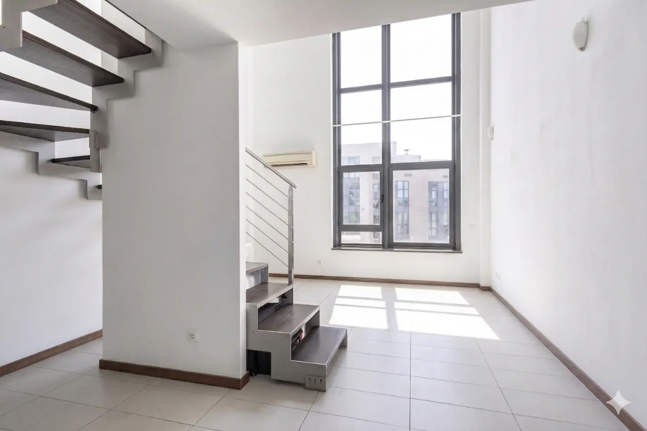 Bright, modern room with white walls, tile floor, and a large window. A dark wood and metal staircase leads up.