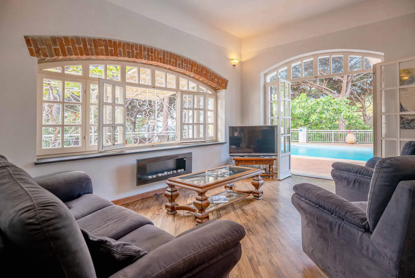 Living room with gray sofas, wood floors, and a glass coffee table. A pool is visible through the open doors.