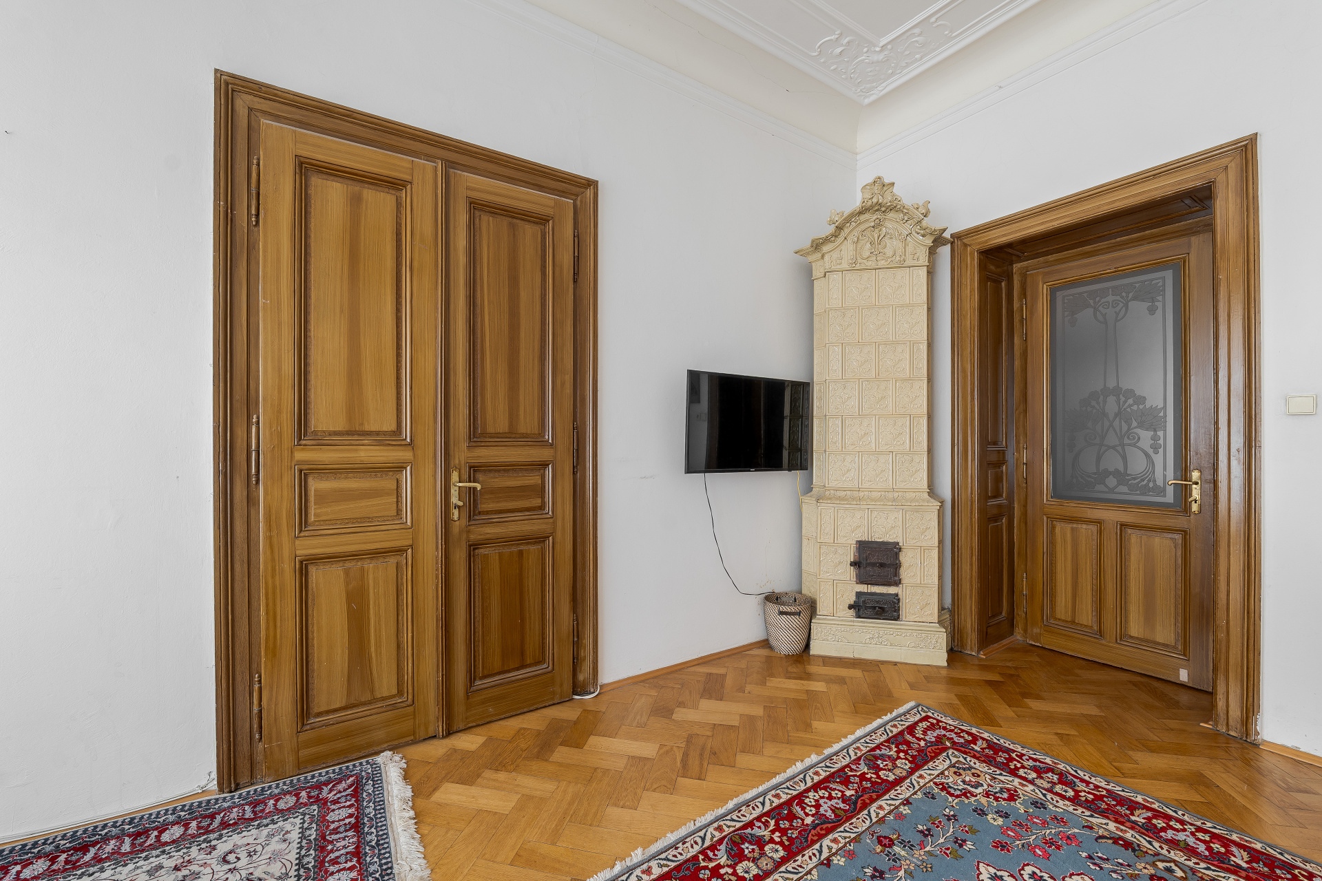 Room with herringbone wood floors, two wooden doors, a vintage tiled stove, and a wall-mounted TV over a small basket. [AI-generated text]
