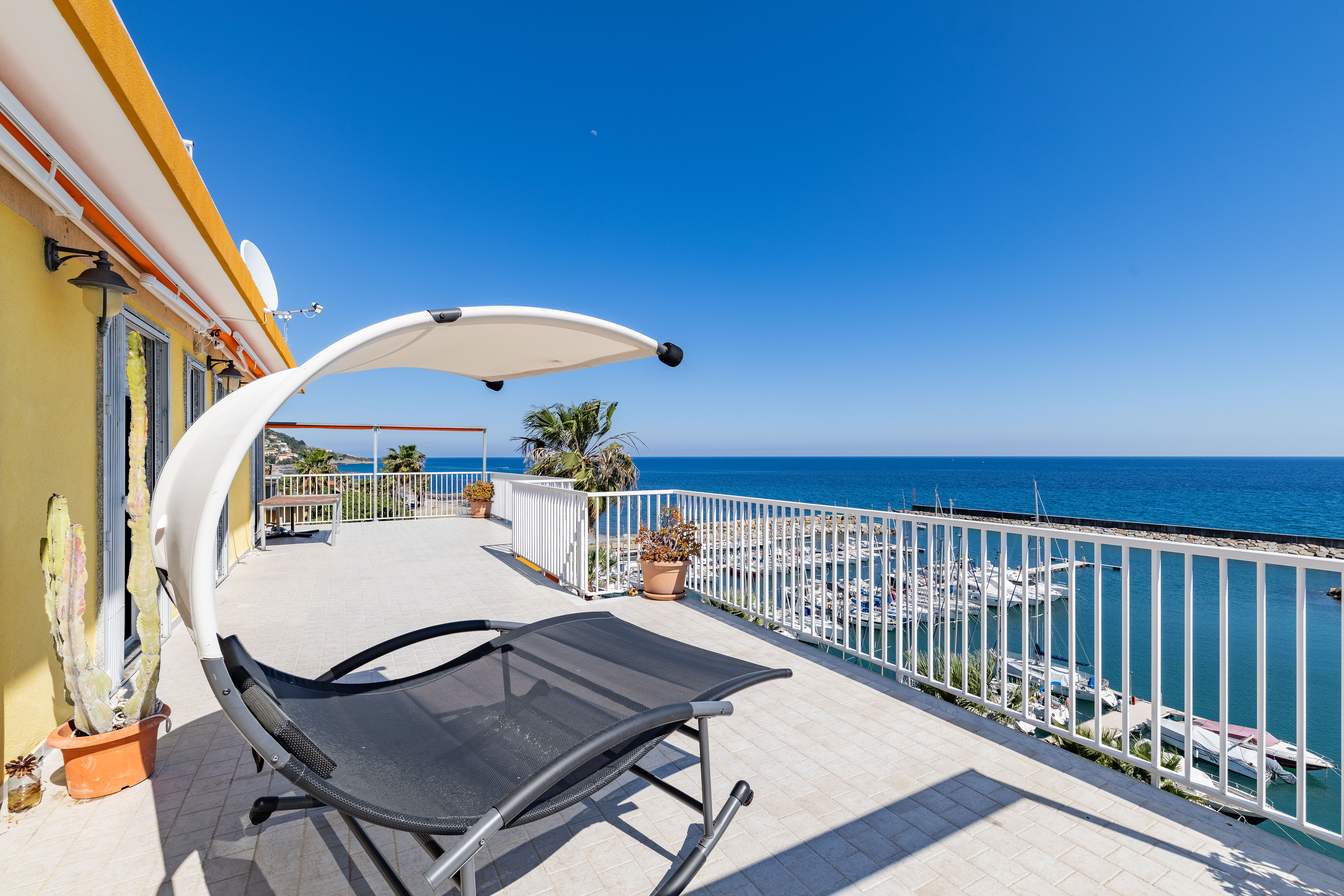 Balcony view of ocean with lounge chair. White railing overlooks marina with boats. Blue sky above. Yellow building on left.