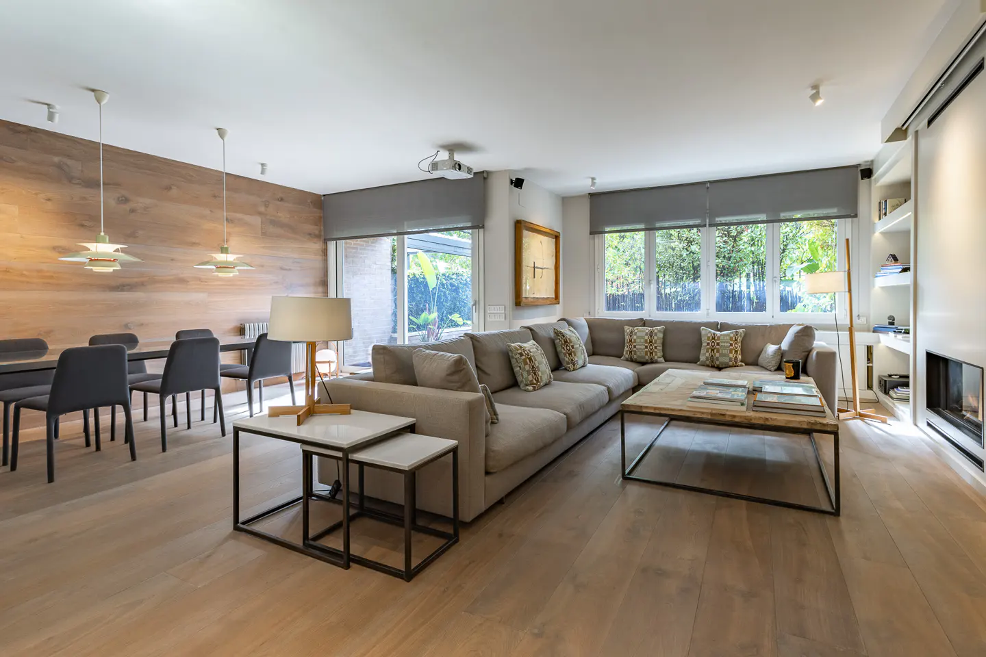 Bright, modern living room with a gray sectional sofa, wood floors, and a dining table with gray chairs. Large windows offer a view of greenery.