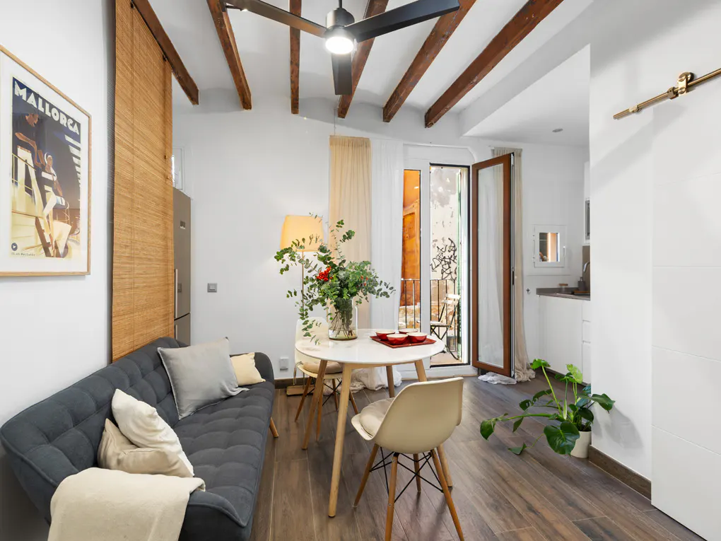 Living room with gray sofa, round white table with flowers, and open balcony door. Ceiling fan and wood beams add character.