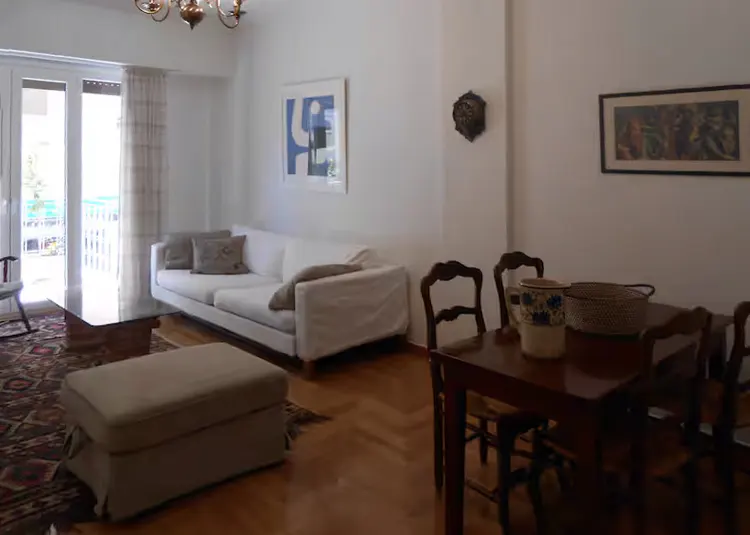 Living room with white sofa, ottoman, and wood dining table with chairs. Art on the white walls and a patterned rug on the wood floor.