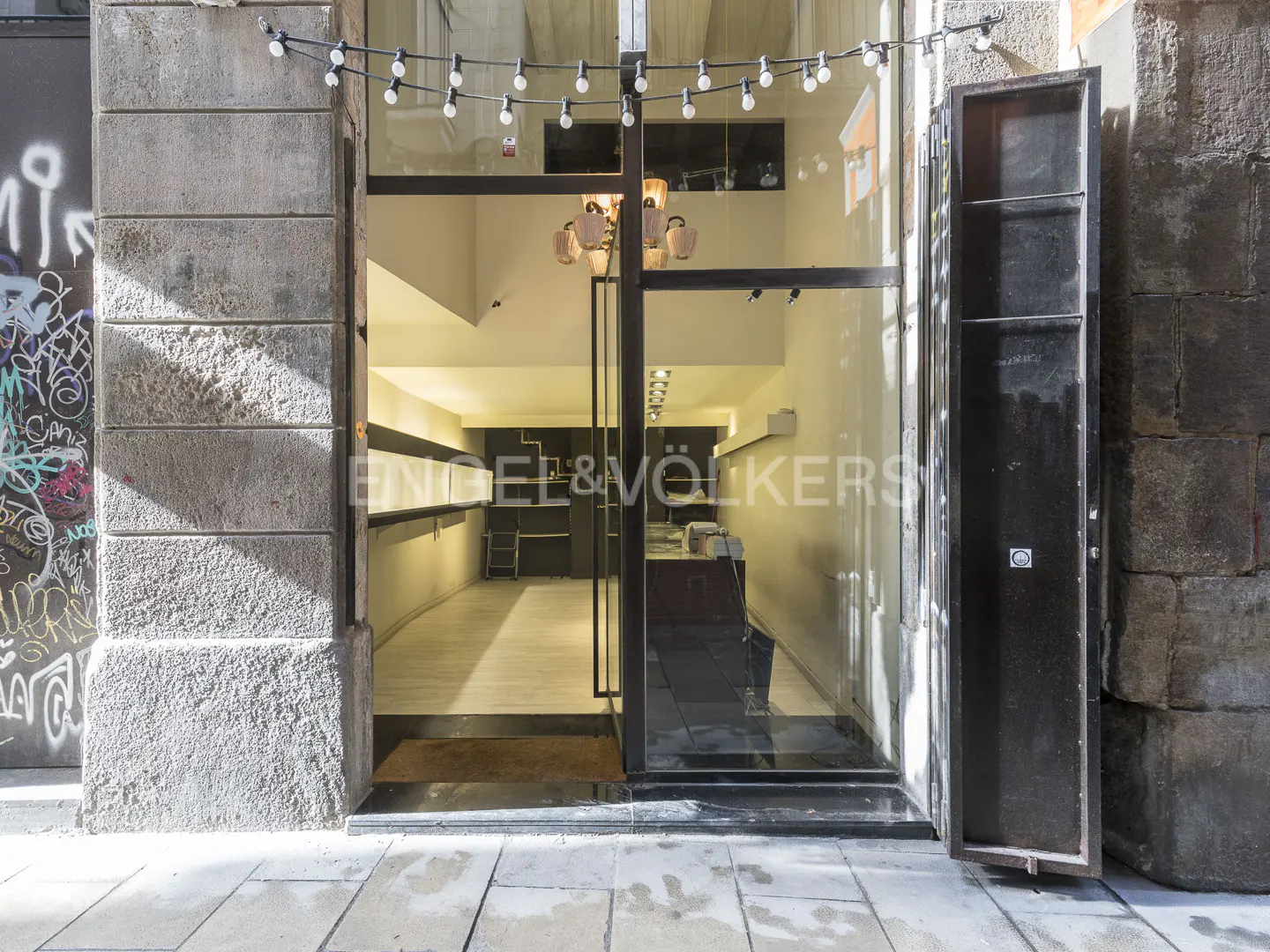 Storefront view with open glass doors, stone walls, and string lights. Engel & Völkers logo visible inside.