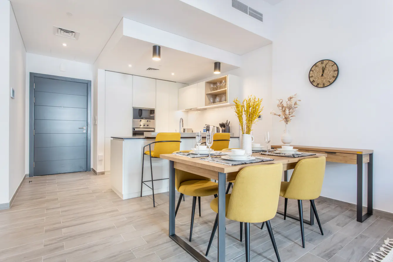Bright, modern apartment interior with a wood dining table set for four, yellow chairs, and a white kitchen in the background.