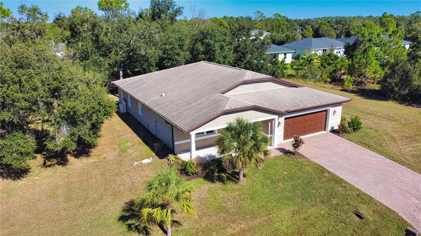 Aerial view of a beige single-story home with a brown roof and garage door, surrounded by green trees and lawn.