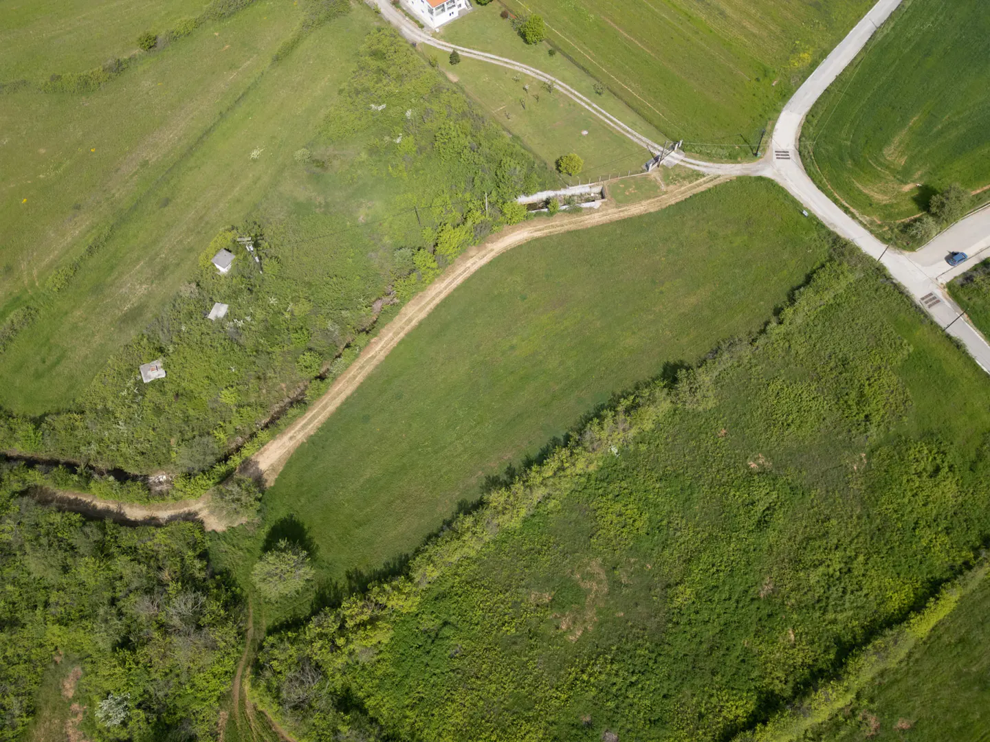 Aerial view of green fields divided by dirt and paved roads, with a white house in the upper left corner.