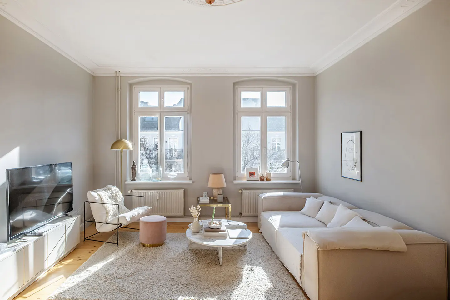 Bright living room with beige sectional sofa, white armchair, and round marble coffee table on a cream shag rug. Two windows let in sunlight.