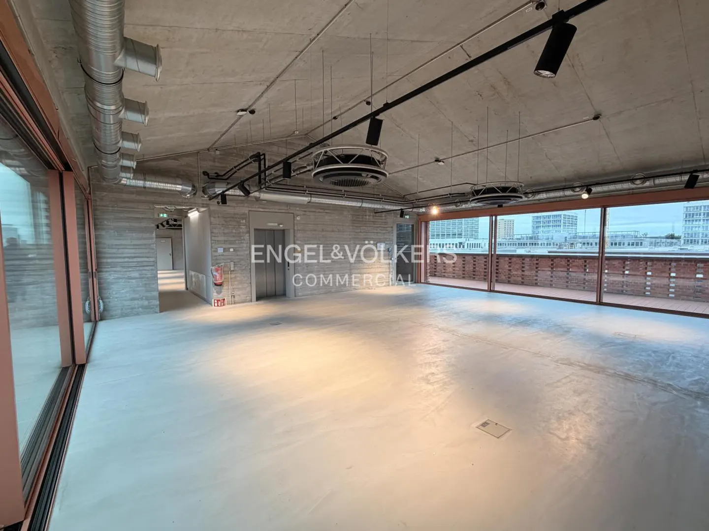 Unfurnished commercial space with concrete floors, walls, and ceiling. Large windows offer city views. Engel & Völkers Commercial logo on the wall.