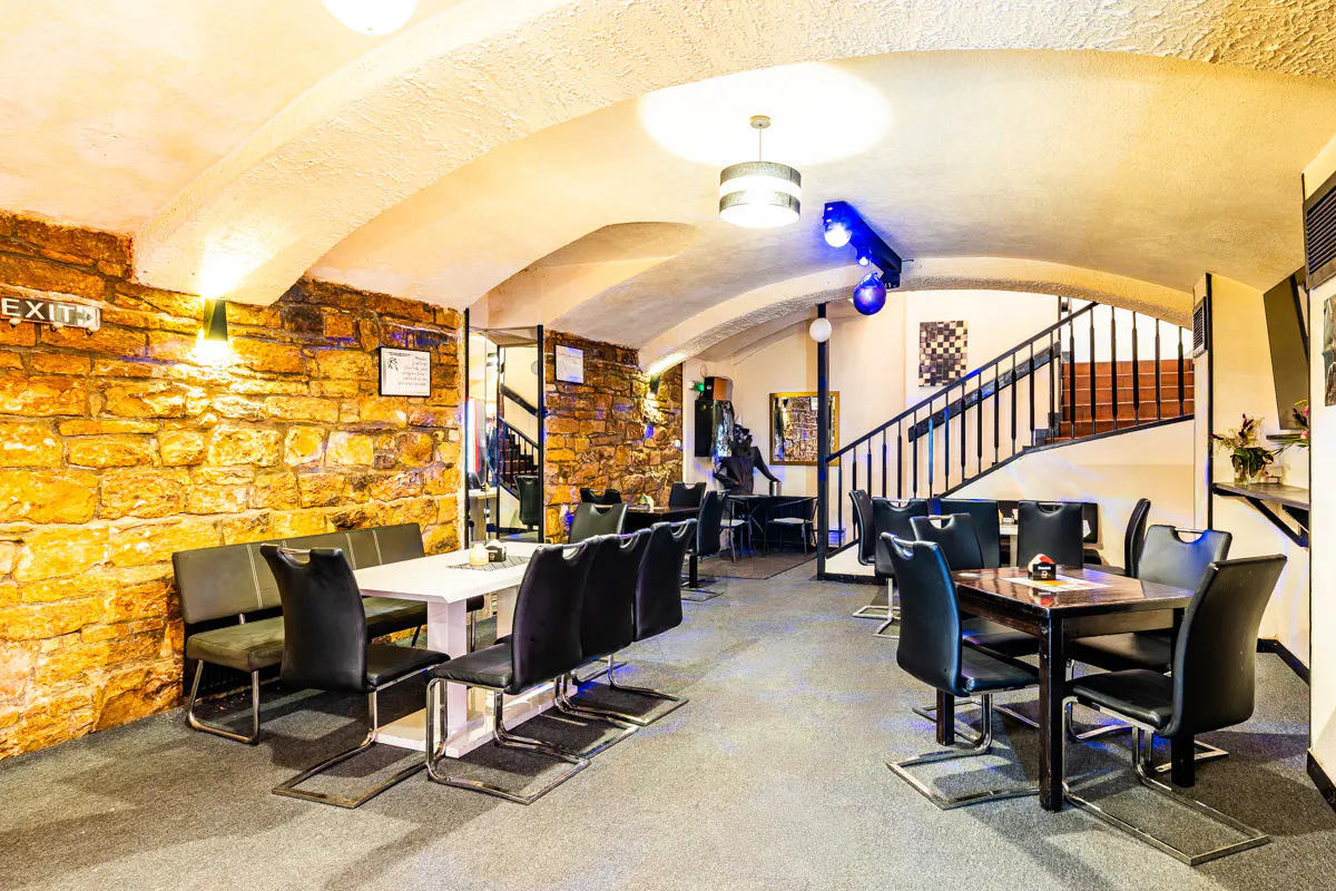 Restaurant interior with stone walls, arched ceilings, black chairs, and tables, featuring a staircase in the background.