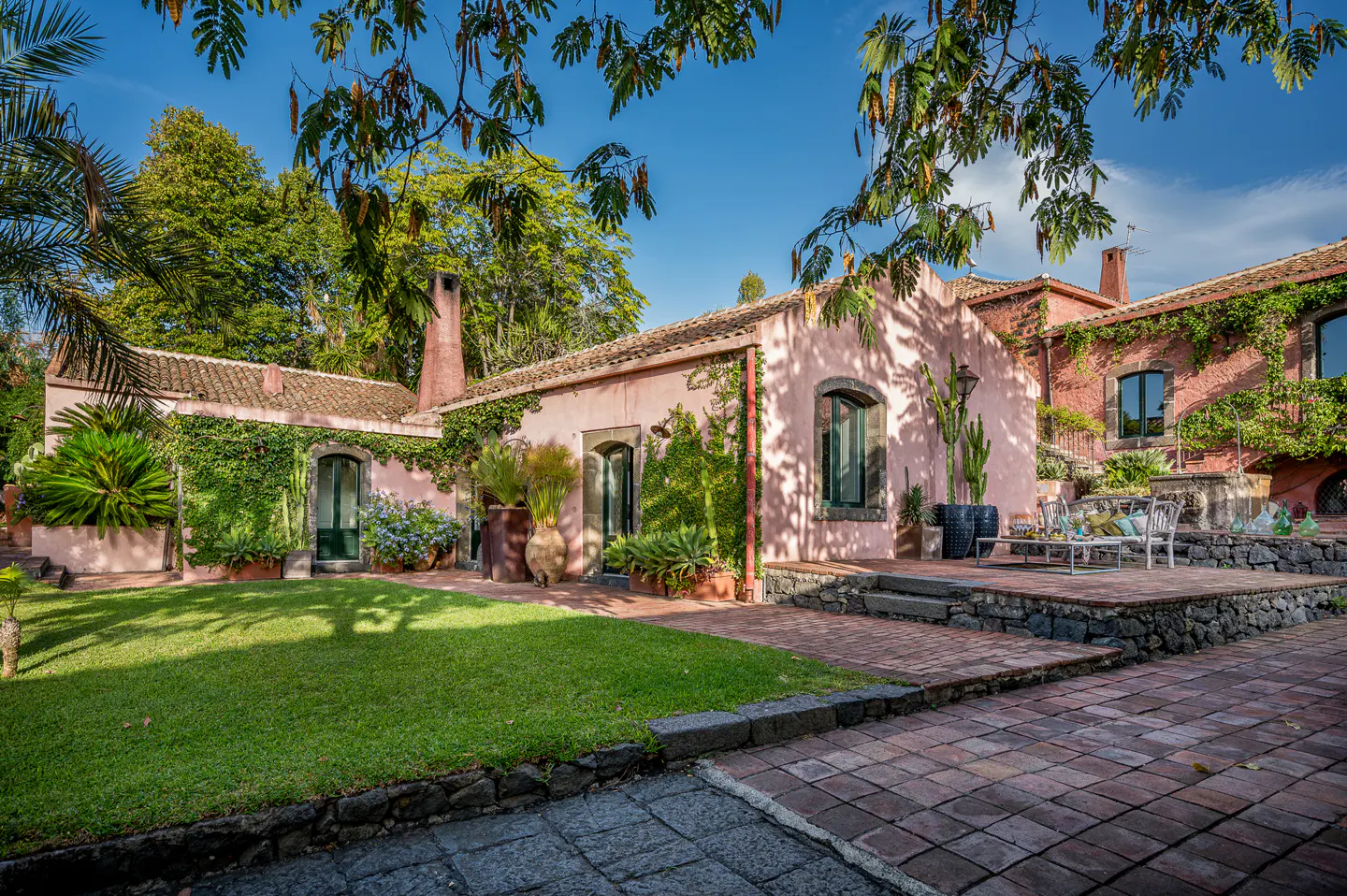 A pink villa with ivy-covered walls, a green lawn, and a brick patio under a bright blue sky.