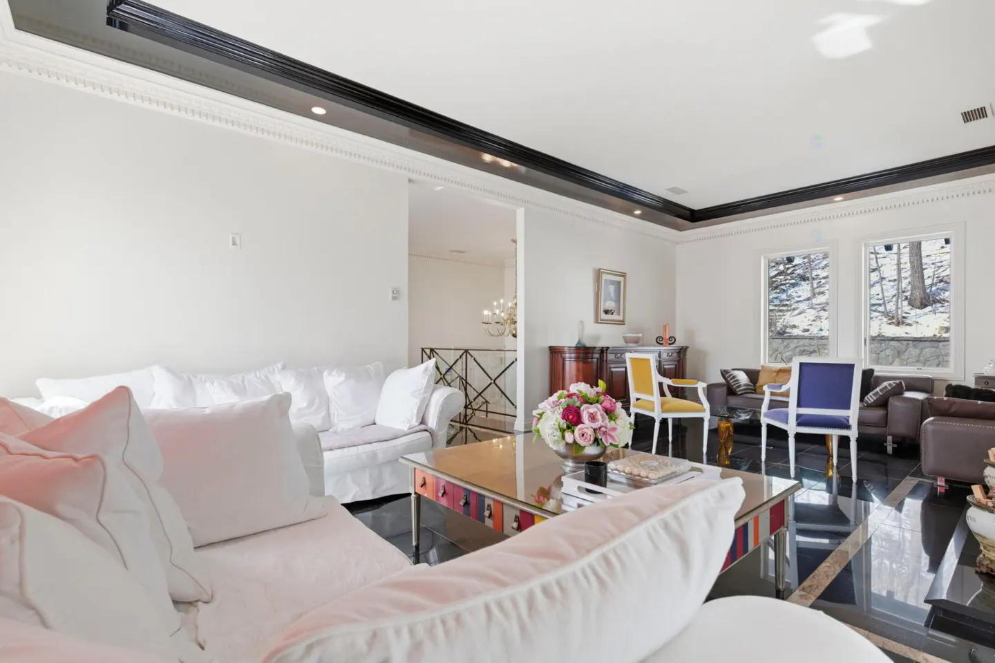 Bright living room with white sofas, a glass coffee table with flowers, and black marble floors. Windows overlook a snowy landscape.