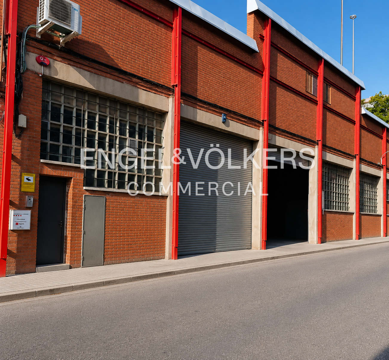Exterior view of a red brick industrial building with large metal roll-up doors, grid windows, and bright red steel beams.