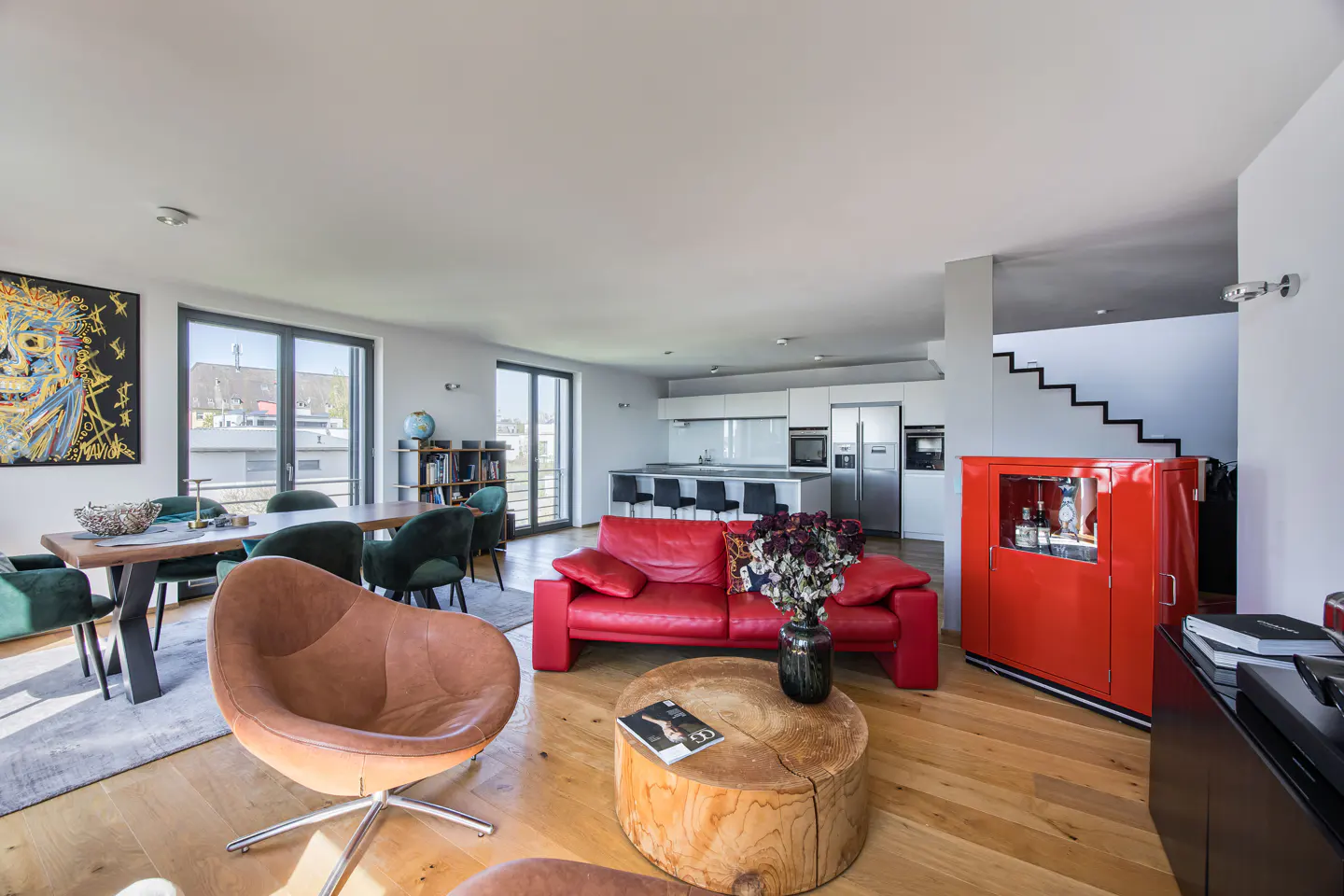 Bright, open-concept living space with wood floors, red sofa, and modern kitchen. Dining table with green chairs and abstract art.