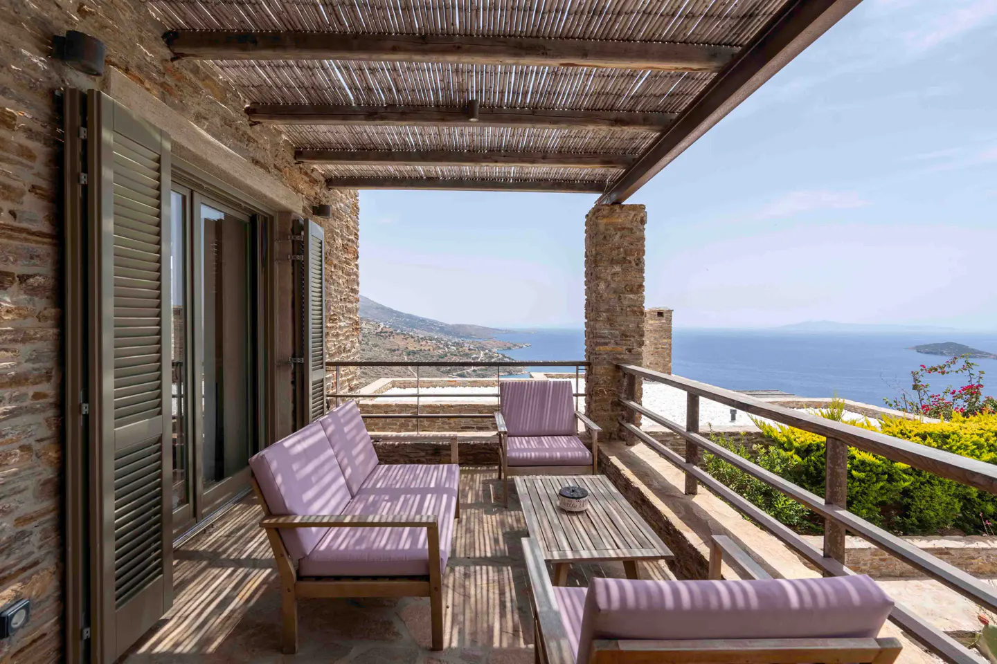 Stone terrace with a wooden pergola, lavender-cushioned seating, and a panoramic view of the blue sea and coastline.