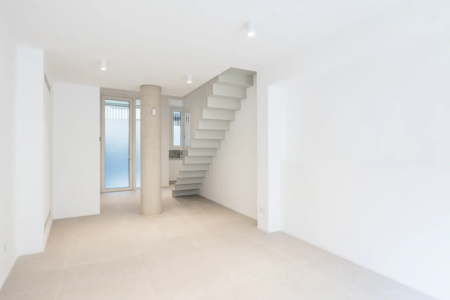 Bright, empty room with white walls, tile floor, and concrete stairs. A concrete pillar stands near a glass door.