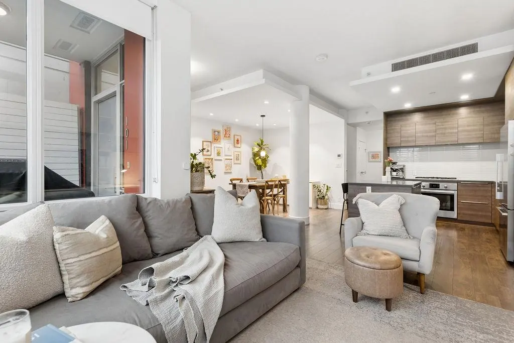 A bright, open-concept living space with a gray sofa, armchair, kitchen, and dining area.