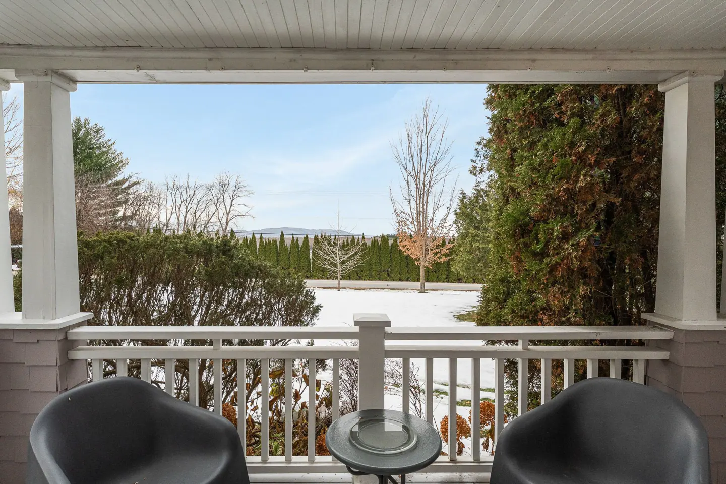 View from a porch with two black chairs and a table, overlooking a snowy yard with trees and a distant body of water.
