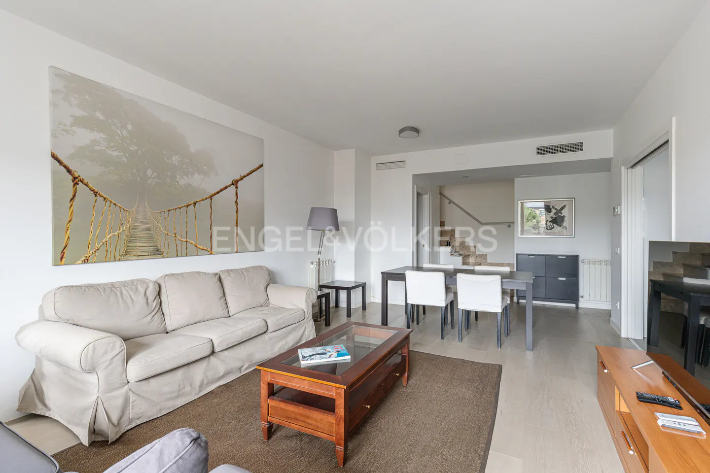 Bright living room with a beige sofa, wooden coffee table, and a large bridge art piece on the wall. Dining table and chairs are in the background.
