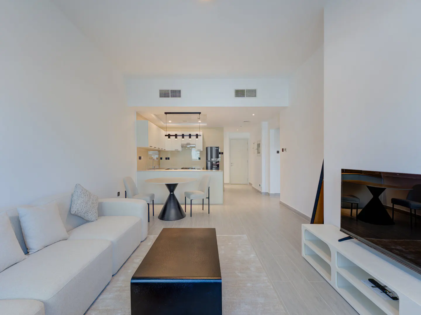 Bright, modern apartment interior with white walls, a white sofa, and a black coffee table. A kitchen and dining area are visible in the background.