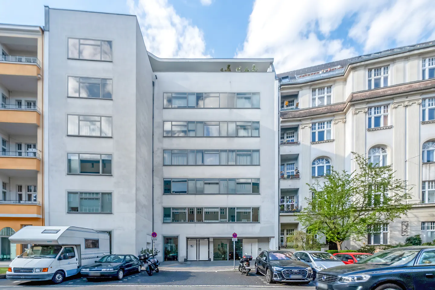 Modern white apartment building facade with large windows, flanked by older buildings, with parked cars and a camper van.