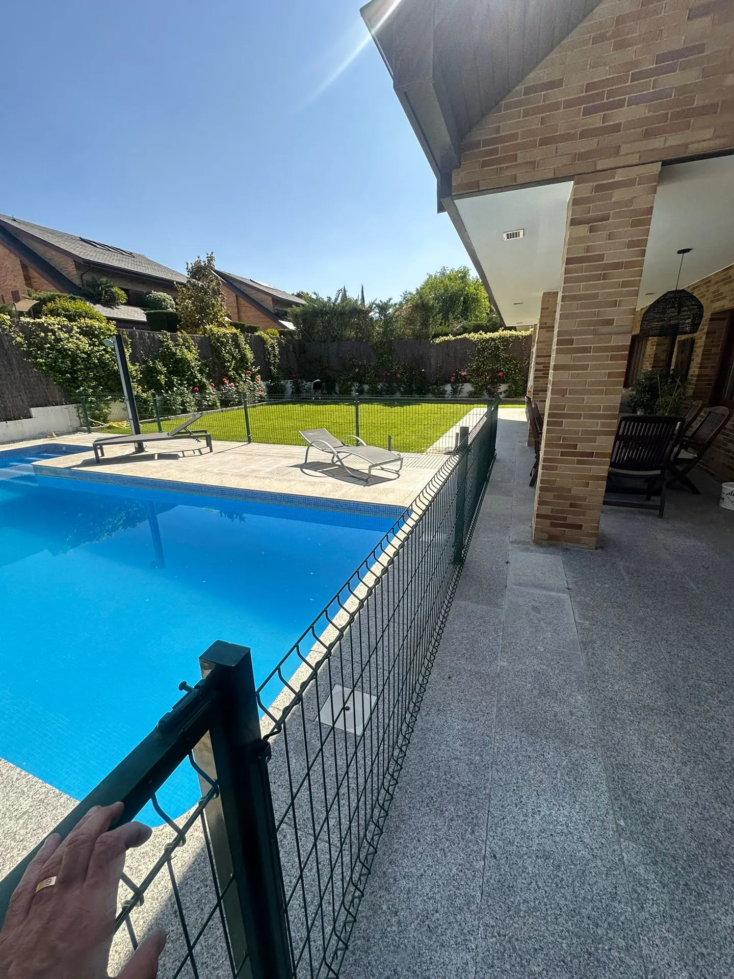 A bright blue swimming pool is enclosed by a black fence, with lounge chairs and green grass visible beyond. A hand rests on the fence.